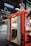 A red food truck with open doors reveals a stainless steel interior, including a sink and shelving. Above, a blue sign reads 'Sägecenter' inside a large industrial warehouse with visible steel beams and pipes. On the right side of the truck, a poster with vivid graphics adds a pop of color.