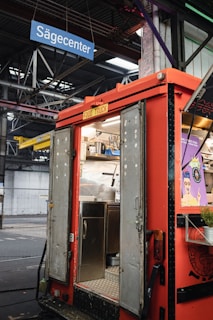 A red food truck with open doors reveals a stainless steel interior, including a sink and shelving. Above, a blue sign reads 'S&auml;gecenter' inside a large industrial warehouse with visible steel beams and pipes. On the right side of the truck, a poster with vivid graphics adds a pop of color.
