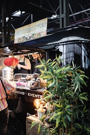 A bustling street food stall named 'Mrs. Chow Chow' offering authentic Asian cuisine. Visible are stacked steamer baskets, a decorative Buddha statue, and vibrant greenery. The warm glow from a hanging red lantern adds an inviting ambiance. People are seen interacting with the vendor inside the stall.