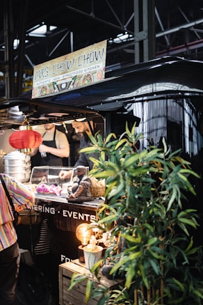 A bustling street food stall named 'Mrs. Chow Chow' offering authentic Asian cuisine. Visible are stacked steamer baskets, a decorative Buddha statue, and vibrant greenery. The warm glow from a hanging red lantern adds an inviting ambiance. People are seen interacting with the vendor inside the stall.