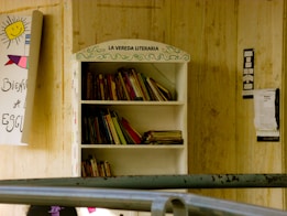 A bookshelf labeled 'La Vereda Literaria' filled with various books, located outdoors against a wooden wall. Nearby, a handmade signs with colorful drawings and text are attached to the wall.