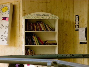 A bookshelf labeled 'La Vereda Literaria' filled with various books, located outdoors against a wooden wall. Nearby, a handmade signs with colorful drawings and text are attached to the wall.