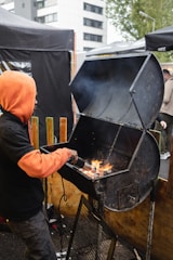 A person is tending to an open barbecue grill with flames visible inside. The individual is wearing a black outfit with an orange hood. The setting appears to be an outdoor area, possibly during a cookout or event, with tents and urban buildings visible in the background.