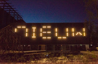 Large illuminated letters spelling 'NIEUW' are mounted on the side of a building at night. The warm glow stands out against the dark, clear night sky. Below, there is a reflective surface that appears to be water, with some vegetation visible around it. The structure has a modern and industrial aesthetic.