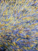 A detailed view of a textured surface composed of intertwined yarns in shades of blue, yellow, and traces of other colors. The fibers are arranged in a chaotic, overlapping manner, creating a visually dynamic pattern.