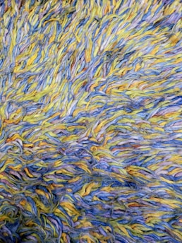 A detailed view of a textured surface composed of intertwined yarns in shades of blue, yellow, and traces of other colors. The fibers are arranged in a chaotic, overlapping manner, creating a visually dynamic pattern.
