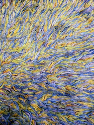 A detailed view of a textured surface composed of intertwined yarns in shades of blue, yellow, and traces of other colors. The fibers are arranged in a chaotic, overlapping manner, creating a visually dynamic pattern.