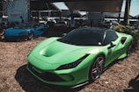 A vibrant green sports car is prominently displayed in the foreground, sitting on a grassy area. In the background, a blue sports car is visible, along with various tents and people gathered around. The setting appears to be a car show or outdoor exhibition, with a banner for 'cleaners studio' visible.