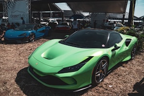 A vibrant green sports car is prominently displayed in the foreground, sitting on a grassy area. In the background, a blue sports car is visible, along with various tents and people gathered around. The setting appears to be a car show or outdoor exhibition, with a banner for 'cleaners studio' visible.