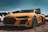 A sleek, yellow sports car with a dynamic design and prominent grille is parked on dry grass. It features intricate wheel designs and bold graphics on the side with the text 'Motor Import.' In the background, other vehicles and tents are partially visible under a clear blue sky.