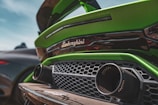 Close-up view of the rear section of a green sports car with the Lamborghini logo prominently displayed. The design features aggressive lines, a black honeycomb grille, and large exhausts, emphasizing the car's high-performance nature.