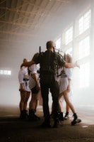 A group of people stands in a large, industrial-style space with high, bright windows. They are wearing coordinated outfits, including shorts, tights, and boots. One person appears to be leading or managing the group, wearing a backpack with equipment and extending their arms towards the others, who are standing in a semi-circle.