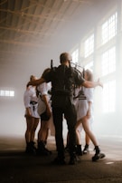A group of people stands in a large, industrial-style space with high, bright windows. They are wearing coordinated outfits, including shorts, tights, and boots. One person appears to be leading or managing the group, wearing a backpack with equipment and extending their arms towards the others, who are standing in a semi-circle.