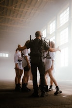 A group of people stands in a large, industrial-style space with high, bright windows. They are wearing coordinated outfits, including shorts, tights, and boots. One person appears to be leading or managing the group, wearing a backpack with equipment and extending their arms towards the others, who are standing in a semi-circle.