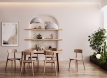 A minimalistic dining area with a wooden table and chairs, two large gray pendant lights, floating wooden shelves decorated with small sculptures and books, and a framed abstract art piece on the wall. A tall green plant is situated on the right, creating a serene and natural ambiance.