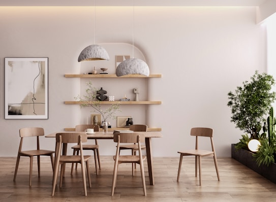 A minimalistic dining area with a wooden table and chairs, two large gray pendant lights, floating wooden shelves decorated with small sculptures and books, and a framed abstract art piece on the wall. A tall green plant is situated on the right, creating a serene and natural ambiance.