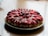 A round strawberry and chocolate dessert arranged on a plate. Sliced strawberries and chocolate pieces are meticulously placed on top of a creamy filling, all resting on a crumbly base.