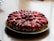 A round strawberry and chocolate dessert arranged on a plate. Sliced strawberries and chocolate pieces are meticulously placed on top of a creamy filling, all resting on a crumbly base.