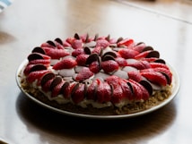 A round strawberry and chocolate dessert arranged on a plate. Sliced strawberries and chocolate pieces are meticulously placed on top of a creamy filling, all resting on a crumbly base.