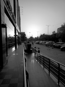 A black and white photograph depicts an urban street scene at dusk or dawn. On the left, a building with reflective windows runs alongside a pathway lined with potted plants and a metal railing. To the right, parked cars are seen along the street, while pedestrians walk in the background. Streetlights are visible against the bright sky, and trees line the distance, contributing to the balanced urban atmosphere.