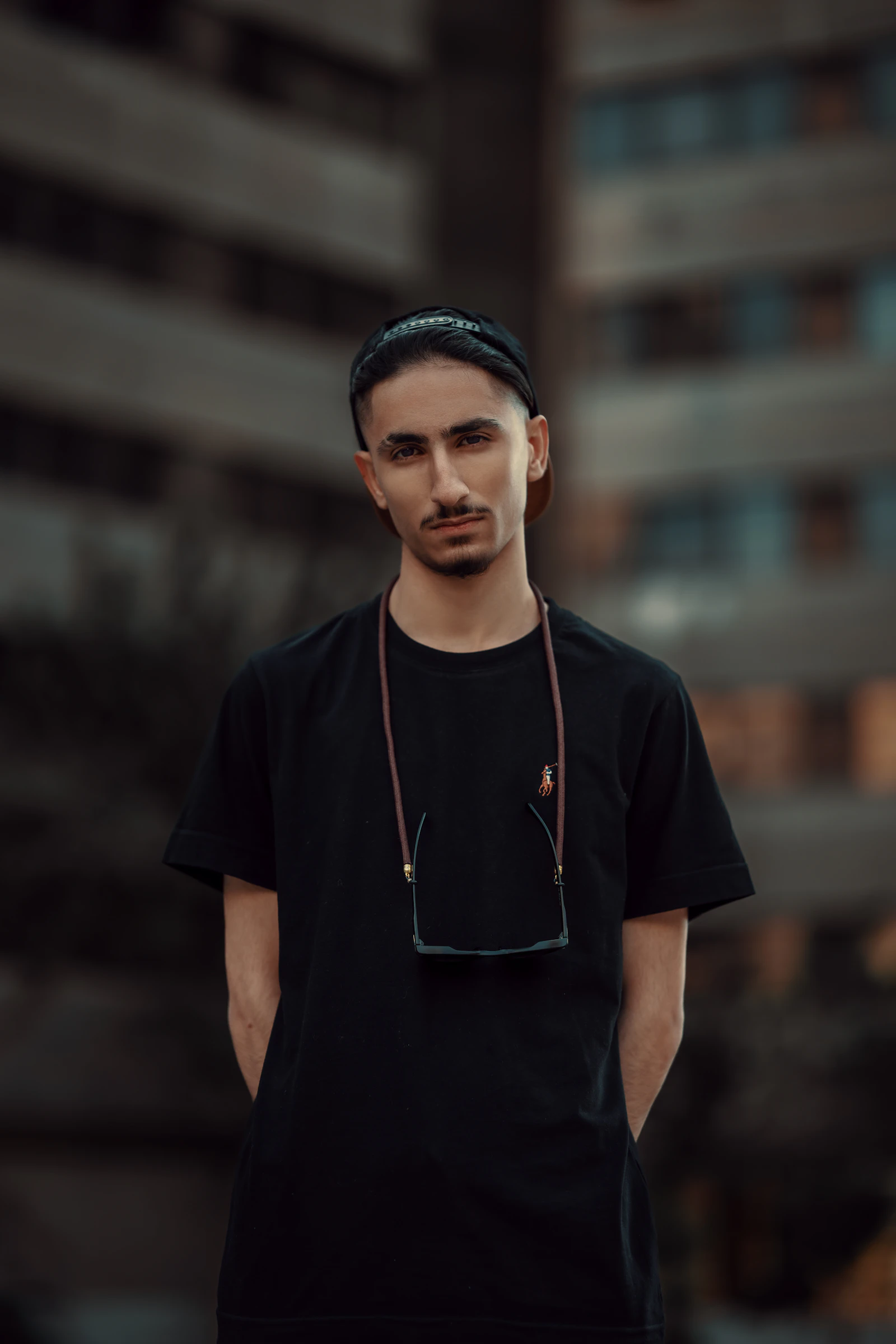 Hombre con camiseta negra y gafas colgadas al cuello, actitud segura, fondo urbano desenfocado