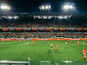A nighttime soccer match taking place in a large stadium filled with a cheering crowd. Players in red and yellow jerseys are actively engaged on the field, with bright floodlights illuminating the scene. The stadium's tiers are packed with enthusiastic fans under a dark sky, and electronic advertisements are visible along the sidelines.