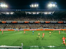 A nighttime soccer match taking place in a large stadium filled with a cheering crowd. Players in red and yellow jerseys are actively engaged on the field, with bright floodlights illuminating the scene. The stadium's tiers are packed with enthusiastic fans under a dark sky, and electronic advertisements are visible along the sidelines.