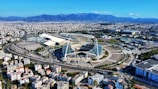 A large, modern sports complex is situated within an expansive urban landscape. The main stadium features prominent architectural structures and an open roof design. Surrounding the stadium are various sports facilities, roads, and greenery. Residential and commercial buildings extend into the distance, framed by mountain ranges under a clear blue sky.