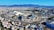 A large, modern sports complex is situated within an expansive urban landscape. The main stadium features prominent architectural structures and an open roof design. Surrounding the stadium are various sports facilities, roads, and greenery. Residential and commercial buildings extend into the distance, framed by mountain ranges under a clear blue sky.