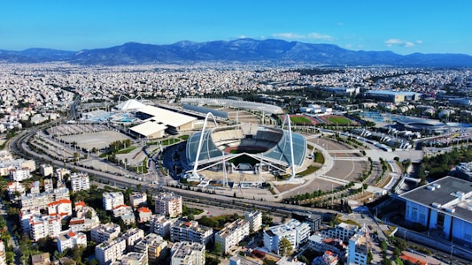 A large, modern sports complex is situated within an expansive urban landscape. The main stadium features prominent architectural structures and an open roof design. Surrounding the stadium are various sports facilities, roads, and greenery. Residential and commercial buildings extend into the distance, framed by mountain ranges under a clear blue sky.