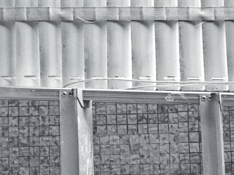 A grayscale image depicting the upper part of a corrugated metal sheet structure, with a patterned band running horizontally across the top portion. The lower part consists of a grid-like design with a mosaic pattern. The combination of textures and patterns suggests an industrial or architectural theme.