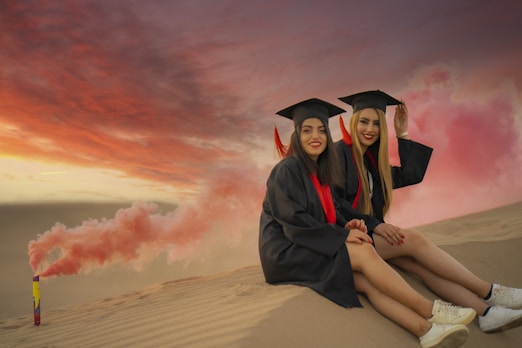two women in graduation gowns sitting on a sand dune