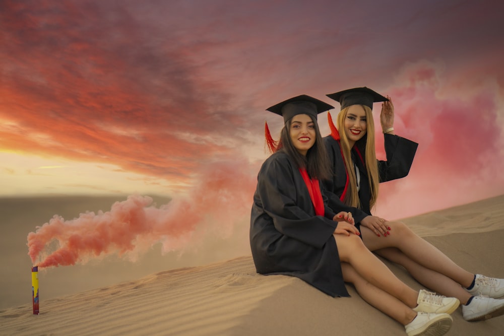 two women in graduation gowns sitting on a sand dune