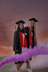 Two women wearing graduation attire, including black gowns, red stoles, and mortarboards, smile happily against a backdrop of a colorful sunset. A vibrant cloud of purple smoke adds dynamic movement to the scene.