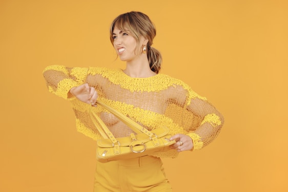 A person wearing a yellow and mesh sweater poses playfully against a matching yellow background, holding a yellow handbag. The scene is vibrant and monochromatic, emphasizing a sense of fun and style.