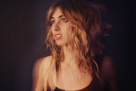 A creative portrait of a woman with long, wavy blonde hair against a dark background. The image has a double exposure effect, with one profile facing forward and another faint profile facing to the right, appearing to shout or express strong emotion.