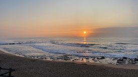 A serene beach scene during sunrise, with the sun partially obscured by clouds on the horizon. Gentle waves roll towards the sandy shore, and a few rocks are scattered along the beach. The sky is painted with soft hues of orange and blue.