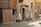 A rustic street scene featuring a carpet gallery with a variety of patterned rugs displayed on the stone wall and ground. Cushions and artwork are neatly arranged outside the shop entrance, creating an inviting and vibrant display.