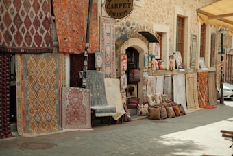 A rustic street scene featuring a carpet gallery with a variety of patterned rugs displayed on the stone wall and ground. Cushions and artwork are neatly arranged outside the shop entrance, creating an inviting and vibrant display.