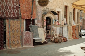 A rustic street scene featuring a carpet gallery with a variety of patterned rugs displayed on the stone wall and ground. Cushions and artwork are neatly arranged outside the shop entrance, creating an inviting and vibrant display.