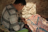 A person in a plaid shirt is engaged in weaving or crafting on a patterned fabric under soft, natural lighting. The background consists of a textured stone wall, and tools like scissors are visible on the fabric.