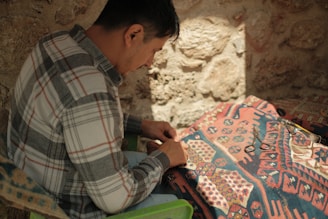 A person in a plaid shirt is engaged in weaving or crafting on a patterned fabric under soft, natural lighting. The background consists of a textured stone wall, and tools like scissors are visible on the fabric.
