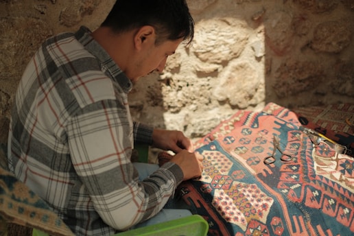 A person in a plaid shirt is engaged in weaving or crafting on a patterned fabric under soft, natural lighting. The background consists of a textured stone wall, and tools like scissors are visible on the fabric.