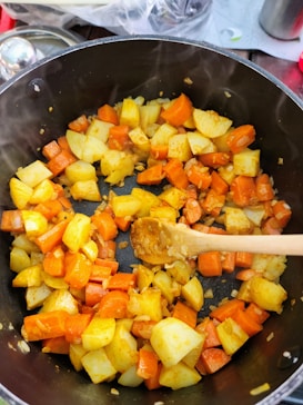 A pot filled with chopped and cooked vegetables, including carrots and potatoes, being stirred with a wooden spoon. There is visible steam indicating that the dish is hot and freshly cooked. The background shows a partially seen kitchen setting.