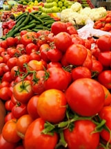 A vibrant display of fresh produce including red tomatoes, green cucumbers, green apples, and white cauliflower, neatly arranged in a market setting. The image highlights the abundance and variety of colorful vegetables and fruits.