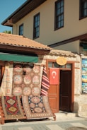 A charming storefront displaying an array of colorful, patterned rugs hangs on a wooden frame outside a stone building. A Turkish flag hangs on a door next to the rugs, and the building features traditional architecture with brown window frames and a terracotta tiled roof.