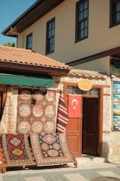 A charming storefront displaying an array of colorful, patterned rugs hangs on a wooden frame outside a stone building. A Turkish flag hangs on a door next to the rugs, and the building features traditional architecture with brown window frames and a terracotta tiled roof.