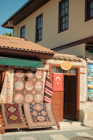 A charming storefront displaying an array of colorful, patterned rugs hangs on a wooden frame outside a stone building. A Turkish flag hangs on a door next to the rugs, and the building features traditional architecture with brown window frames and a terracotta tiled roof.