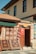 A charming storefront displaying an array of colorful, patterned rugs hangs on a wooden frame outside a stone building. A Turkish flag hangs on a door next to the rugs, and the building features traditional architecture with brown window frames and a terracotta tiled roof.