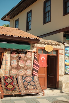 A charming storefront displaying an array of colorful, patterned rugs hangs on a wooden frame outside a stone building. A Turkish flag hangs on a door next to the rugs, and the building features traditional architecture with brown window frames and a terracotta tiled roof.